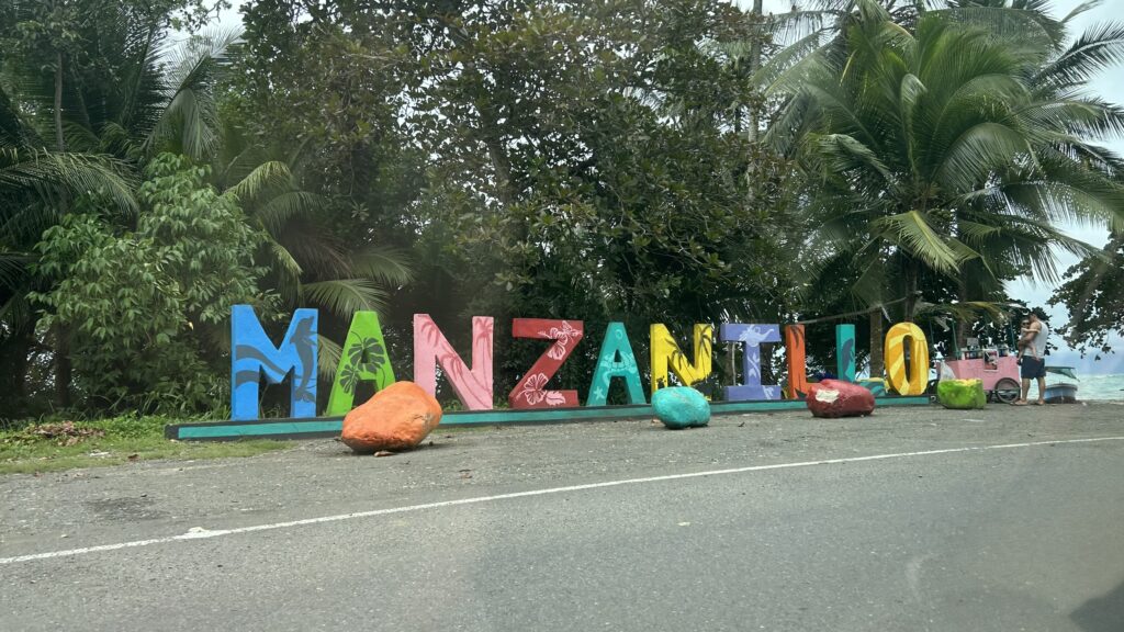Colorful "Manzanillo" sign along a roadside surrounded by lush palm trees and greenery.
