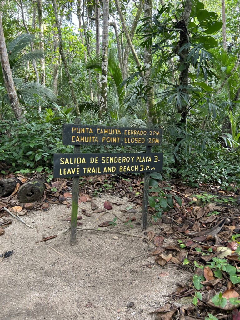 A wooden sign in a tropical forest displaying trail and beach closure times in English and Spanish.